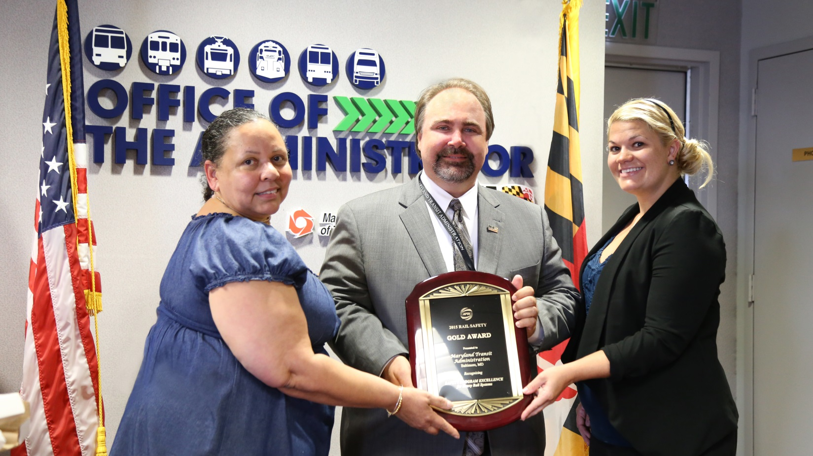 MTA Administrator Paul Comfort displays the Gold Award for Safety that MTA earned from the American Public Transportation Association for the agency&rsquo;s Safety Rules and Compliance Program (SRCP), designed to keep customers, employees and equipment safe. MTA's Bernadette Bridges, head of the Office of Safety, Quality Assurance and Risk Management shares in the honor, left, assisted by Transportation Planning consultant Kimiya Rae Aghevli Darrell, right.