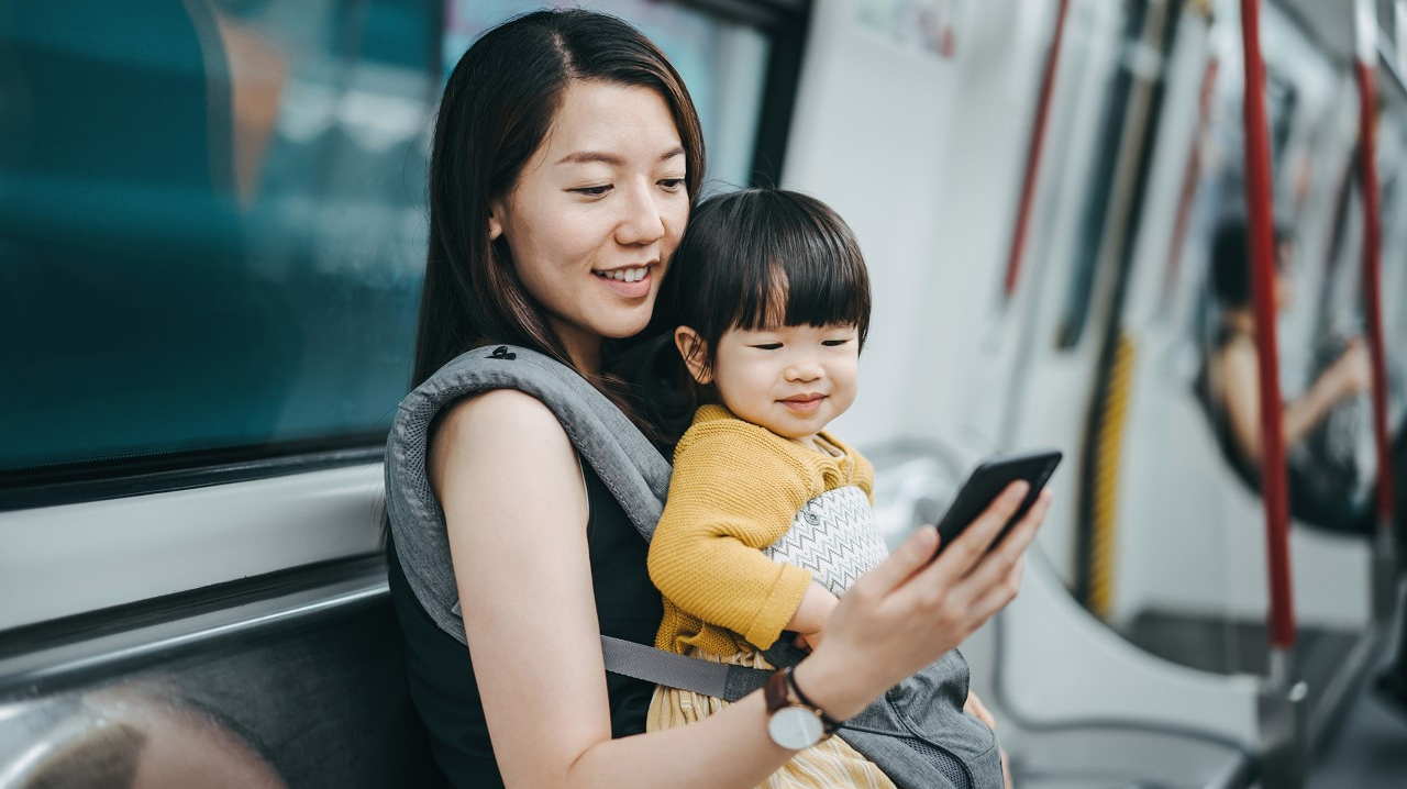 A mom traveling while holding her baby and looking at her phone.