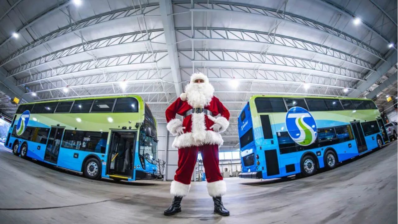 person dressed as santa stands between two double decker buses inside bus garage