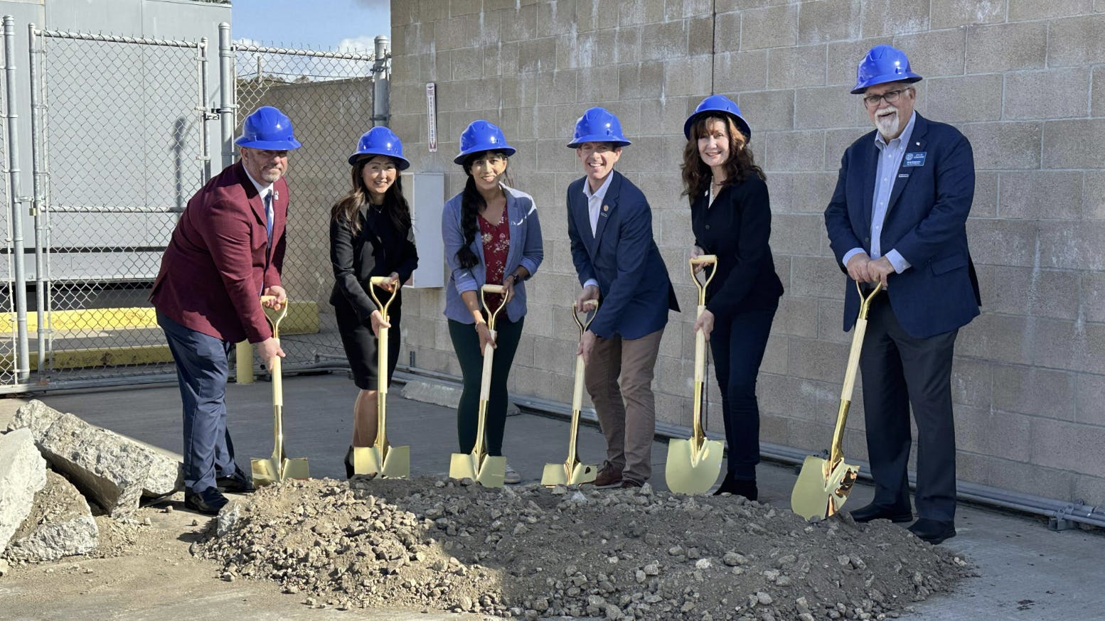 People stand in a line with shovels in a pile of dirt and blue hard hats on heads