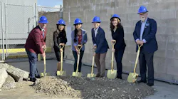 People stand in a line with shovels in a pile of dirt and blue hard hats on heads