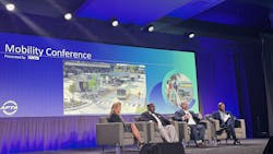 four speakers sit in a line in chairs on a stage with mobility conference backdrop four speakers sit in a line in chairs on a stage with mobility conference backdrop