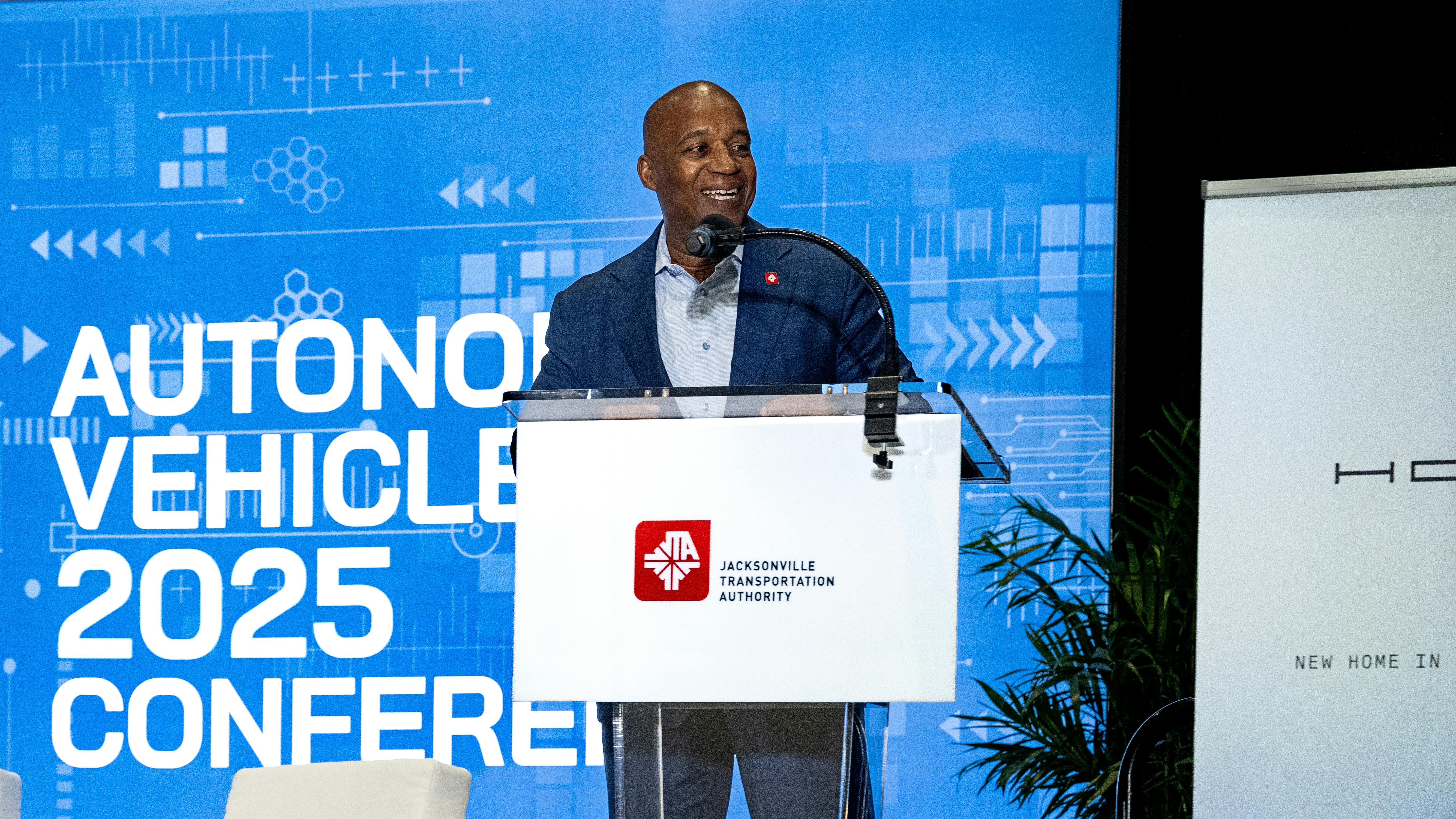 JTA CEO Nat Ford stands at a podium with the JTA logo on the front and a screen behind him that says autonomous vehicle 2025 conference