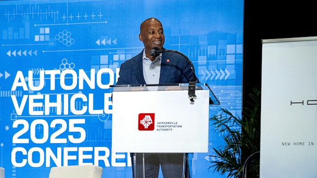 JTA CEO Nat Ford stands at a podium with the JTA logo on the front and a screen behind him that says autonomous vehicle 2025 conference