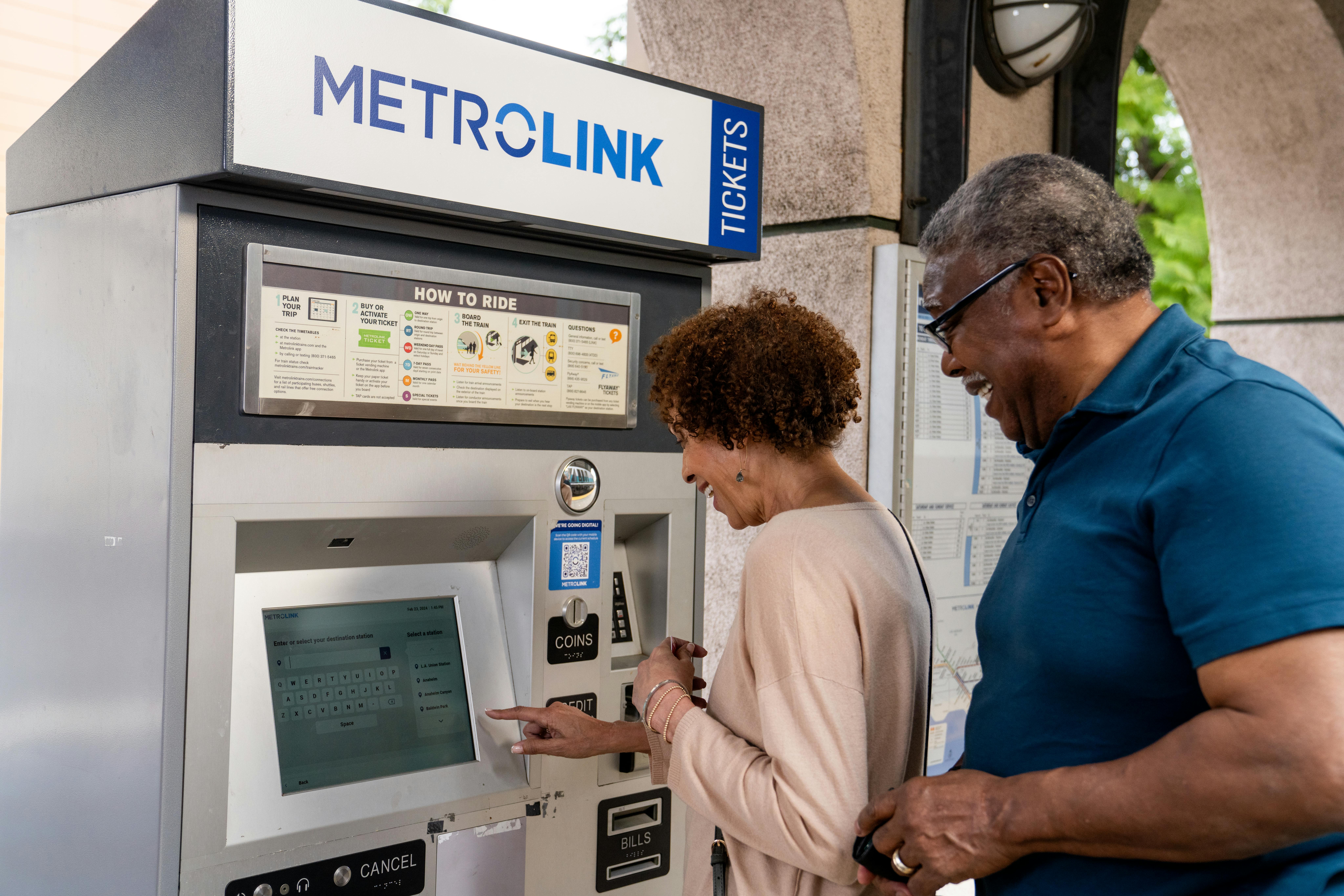 a man and a woman stand in front of a Metrolink ticket vending machine to purchase a ticket