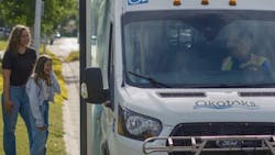 a woman and girl stand on a curb next to an Okotoks Transit cutaway bus a woman and girl stand on a curb next to an Okotoks Transit cutaway bus