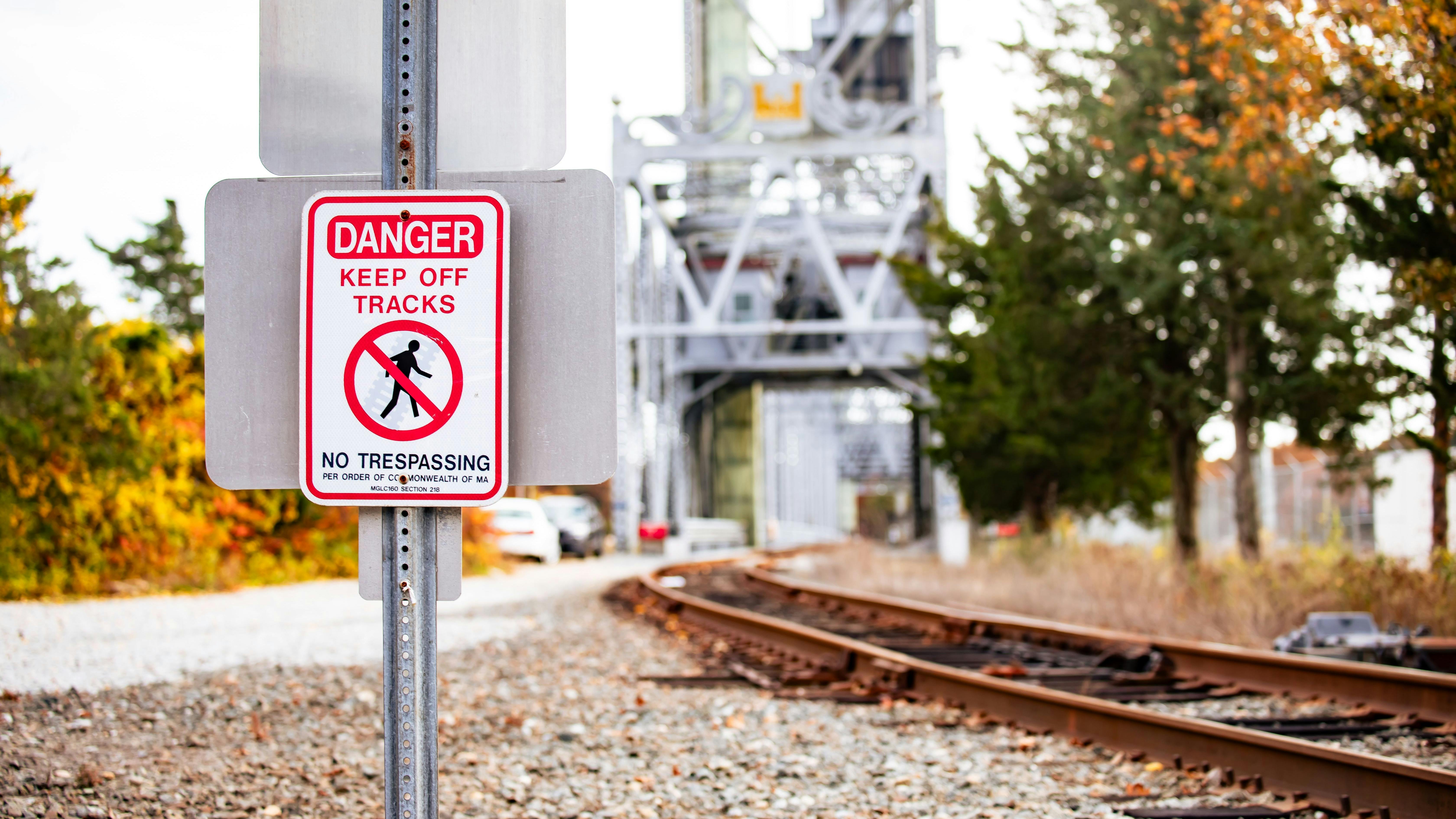 Keep Off Tracks Warning. A warning sign reading Danger Keep Off Tracks stands beside a railway, with autumn trees and the Cape Cod Canal Railroad Bridge in the background.