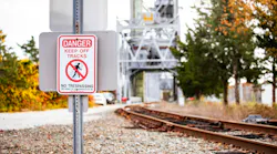 Keep Off Tracks Warning. A warning sign reading Danger Keep Off Tracks stands beside a railway, with autumn trees and the Cape Cod Canal Railroad Bridge in the background.