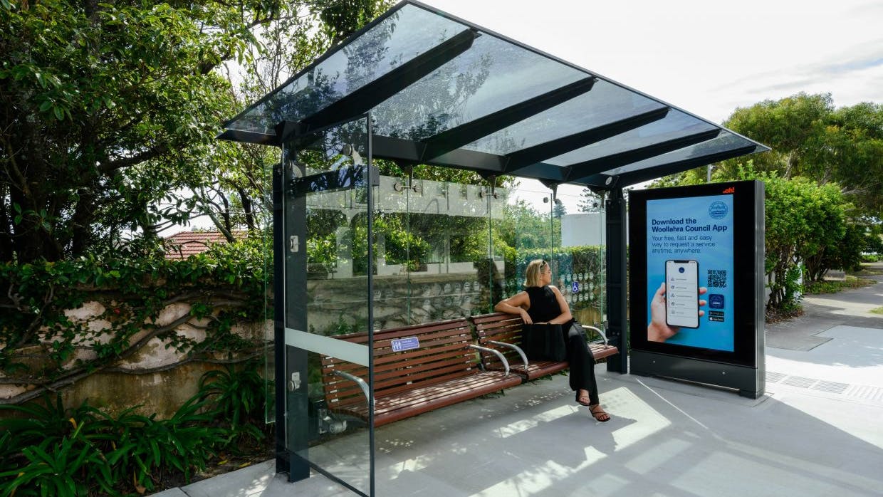 The image shows a woman sitting on a bus stop bench looking at an ad on a Praevar Podium display.