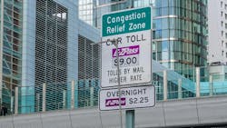 The image shows a Congestion Relief Zone pricing sign hanging above a toll booth. The image shows a Congestion Relief Zone pricing sign hanging above a toll booth.