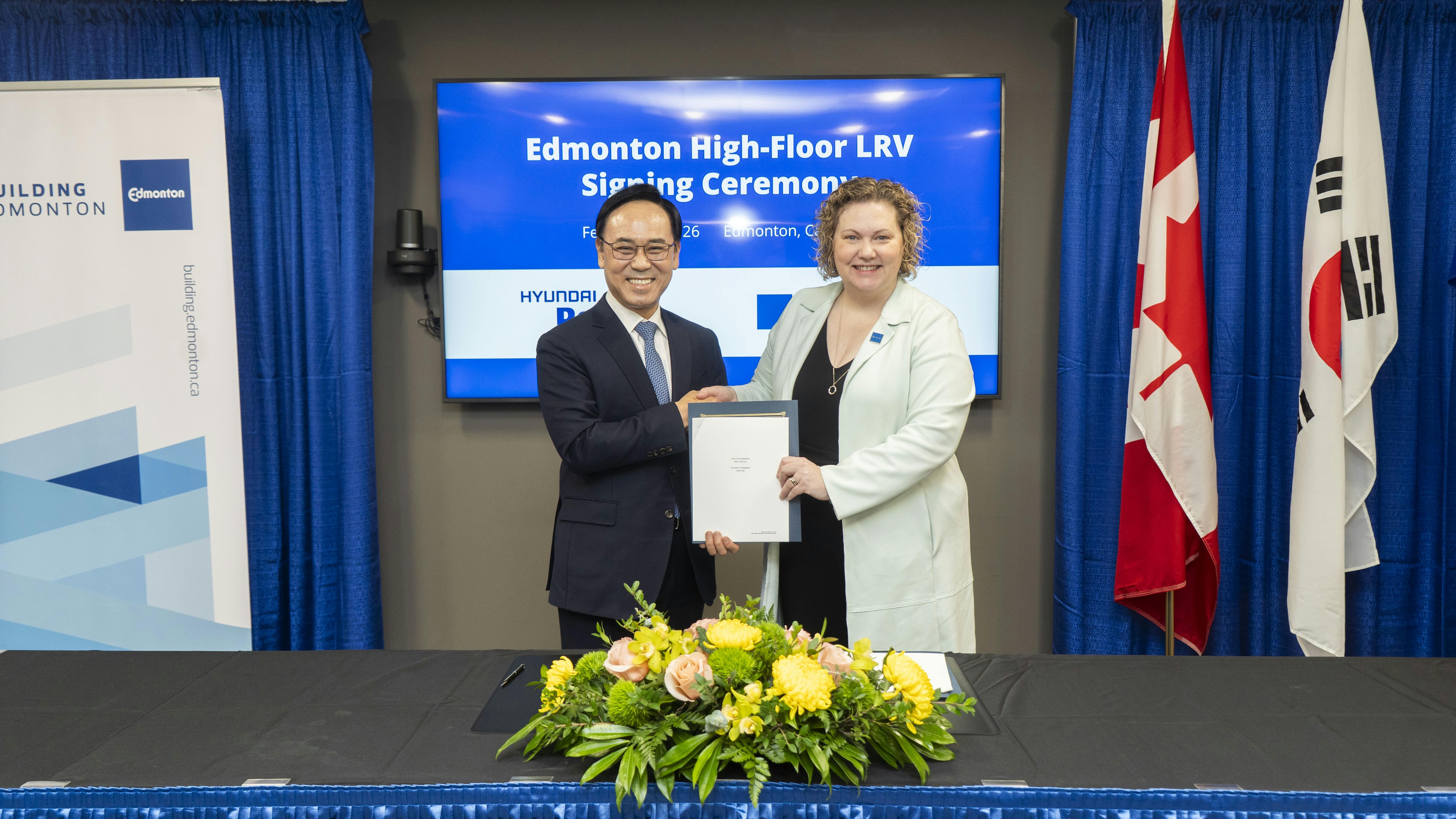 A leader from each the city of Edmonton and HRC hold up the newly signed contract in front of an event marquee.