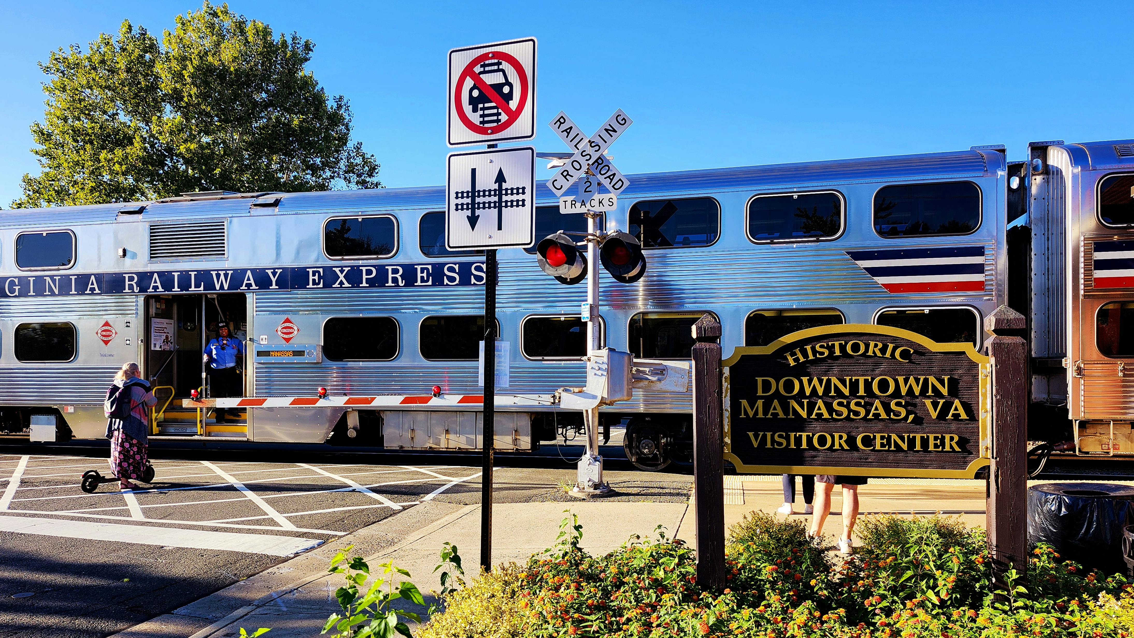 A VRE train passes a crossing in front of a waiting scooter rider.