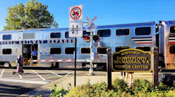 A VRE train passes a crossing in front of a waiting scooter rider.