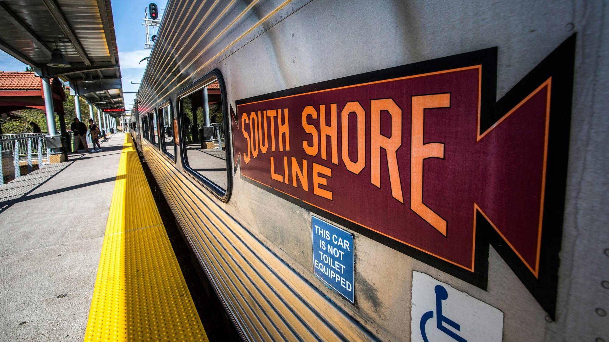 A Northern Indiana Commuter Transportation District South Shore Line commuter rail car sits at a station platform. The stainless steel car bears a maroon and orange 'South Shore Line' herald. A blue placard reads 'This car is not toilet equipped,' and an accessibility symbol is visible below it. The yellow tactile warning strip lines the platform edge.