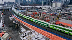 An aerial view of a GO Transit bi-level commuter rail train passing through an active construction zone in winter. The green and white train travels along a corridor flanked by orange safety fencing, construction equipment, cranes, and building materials. The Toronto skyline, including the CN Tower, is visible in the background.
