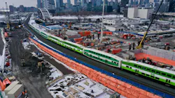 An aerial view of a GO Transit bi-level commuter rail train passing through an active construction zone in winter. The green and white train travels along a corridor flanked by orange safety fencing, construction equipment, cranes, and building materials. The Toronto skyline, including the CN Tower, is visible in the background. An aerial view of a GO Transit bi-level commuter rail train passing through an active construction zone in winter. The green and white train travels along a corridor flanked by orange safety fencing, construction equipment, cranes, and building materials. The Toronto skyline, including the CN Tower, is visible in the background.