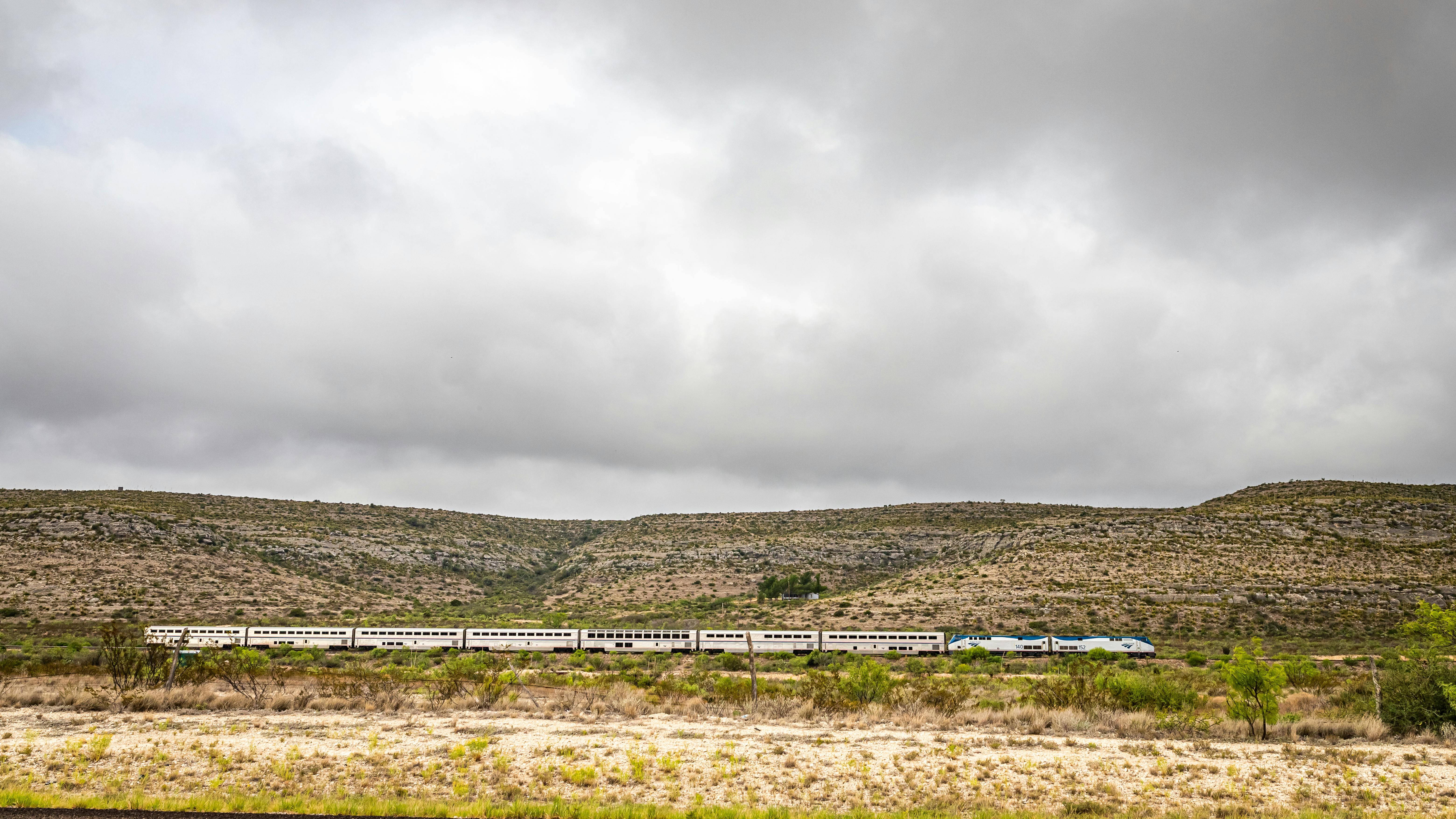 A Sunset Limited train crosses a remote, arid landscape under a heavily overcast sky. The long consist of white bi-level Superliner cars, led by an Amtrak locomotive in the phase VI paint scheme, stretches across the middle distance against scrub-covered hills. Sparse desert vegetation fills the foreground.