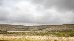 A Sunset Limited train crosses a remote, arid landscape under a heavily overcast sky. The long consist of white bi-level Superliner cars, led by an Amtrak locomotive in the phase VI paint scheme, stretches across the middle distance against scrub-covered hills. Sparse desert vegetation fills the foreground.