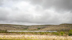 A Sunset Limited train crosses a remote, arid landscape under a heavily overcast sky. The long consist of white bi-level Superliner cars, led by an Amtrak locomotive in the phase VI paint scheme, stretches across the middle distance against scrub-covered hills. Sparse desert vegetation fills the foreground. A Sunset Limited train crosses a remote, arid landscape under a heavily overcast sky. The long consist of white bi-level Superliner cars, led by an Amtrak locomotive in the phase VI paint scheme, stretches across the middle distance against scrub-covered hills. Sparse desert vegetation fills the foreground.