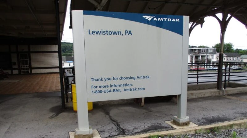 An Amtrak station sign at Lewistown, Pennsylvania, reading 'Thank you for choosing Amtrak. For more information: 1-800-USA-RAIL Amtrak.com.' The sign stands on a platform under a covered canopy, with a small-town streetscape visible in the background.