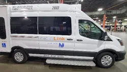 A New Jersey Transit MicroLink van, vehicle number 25801, photographed inside a maintenance facility. The white Ford Transit-based cutaway van displays the MicroLink branding and NJ Transit logo on its side, along with an accessibility symbol indicating wheelchair accessibility. A fold-out entry step is visible below the side door. A New Jersey Transit MicroLink van, vehicle number 25801, photographed inside a maintenance facility. The white Ford Transit-based cutaway van displays the MicroLink branding and NJ Transit logo on its side, along with an accessibility symbol indicating wheelchair accessibility. A fold-out entry step is visible below the side door.