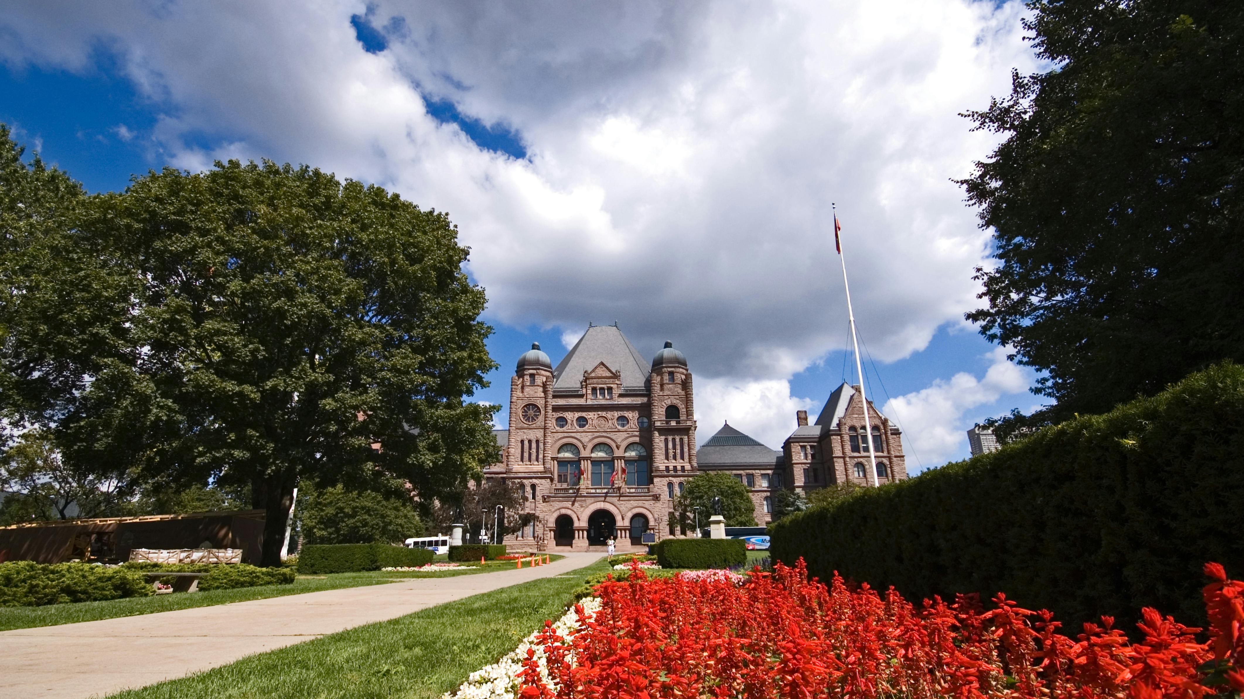 The Ontario Legislative Assembly building at Queen's Park in Toronto, viewed from the front grounds on a sunny summer day. The Romanesque Revival sandstone building is framed by mature trees and a foreground of red and white flowering gardens. A Canadian flag flies from a flagpole to the right of the building.