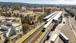 Aerial view of Fruitvale BART Station in Oakland, California. A yellow-striped BART train is stopped at the elevated station platform, which is flanked on the left by the Fruitvale Transit Village — a mixed-use development featuring ground-floor retail, pedestrian plazas, and palm trees. To the right, AC Transit buses are staged in a dedicated bus transfer facility. Residential hills are visible in the background. Aerial view of Fruitvale BART Station in Oakland, California. A yellow-striped BART train is stopped at the elevated station platform, which is flanked on the left by the Fruitvale Transit Village — a mixed-use development featuring ground-floor retail, pedestrian plazas, and palm trees. To the right, AC Transit buses are staged in a dedicated bus transfer facility. Residential hills are visible in the background.