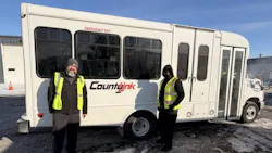 Two transit workers wearing yellow high-visibility vests and winter jackets stand on either side of a white small-transit bus branded with the CountyLink logo. Light snow covers the ground, and a garage or depot building is visible in the background, suggesting a cold-weather operating environment. Two transit workers wearing yellow high-visibility vests and winter jackets stand on either side of a white small-transit bus branded with the CountyLink logo. Light snow covers the ground, and a garage or depot building is visible in the background, suggesting a cold-weather operating environment.