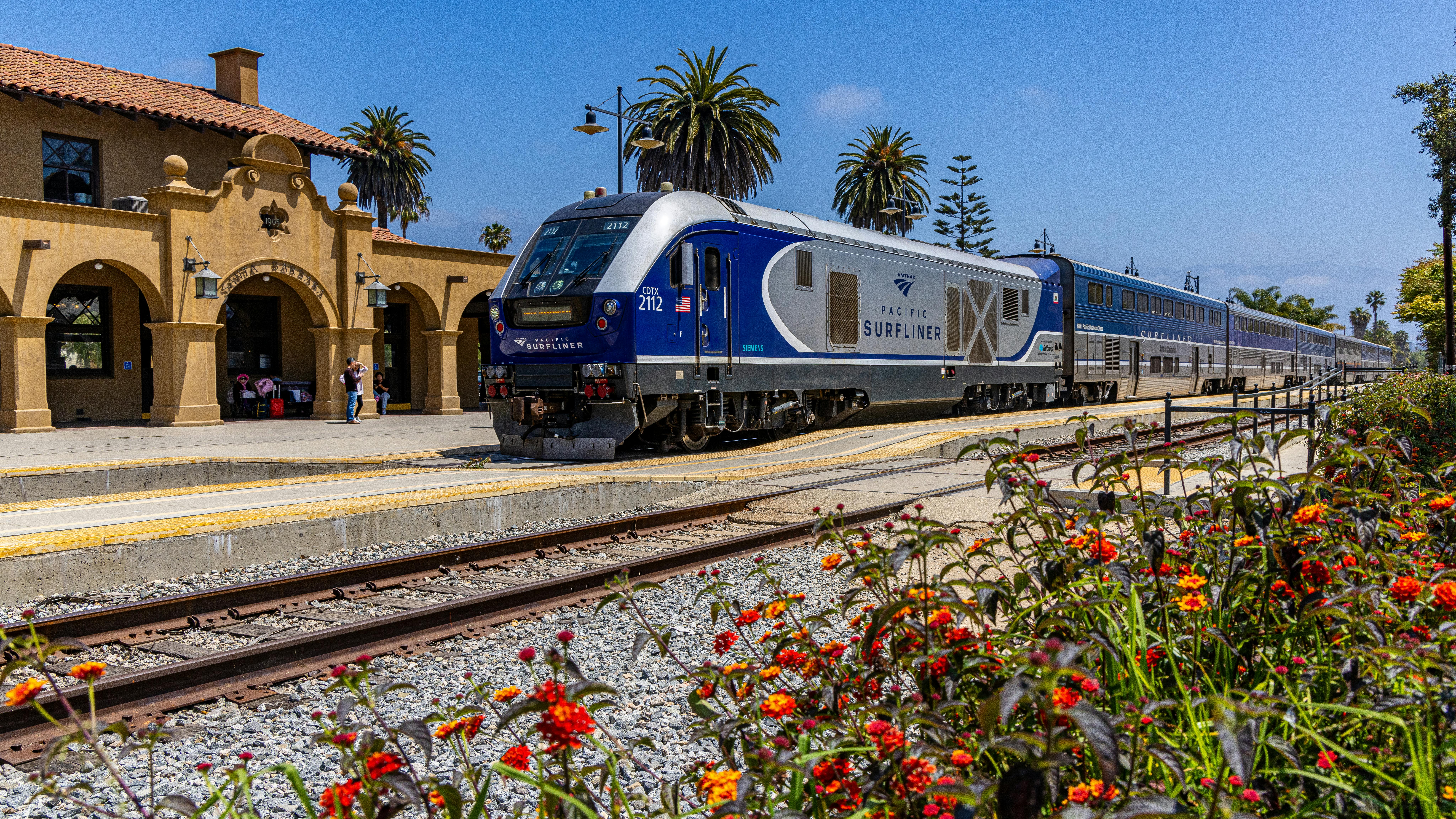 Pacific Surfliner train No. 2112 arrives at Santa Barbara station on a clear day. The blue-and-silver Siemens-built locomotive leads a consist of bilevel passenger cars along the platform of the station's Spanish Colonial Revival depot, its tan stucco walls and red tile roof visible at left. Red and orange lantana blooms fill the foreground, with palm trees and a deep blue sky in the background.