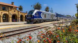 Pacific Surfliner train No. 2112 arrives at Santa Barbara station on a clear day. The blue-and-silver Siemens-built locomotive leads a consist of bilevel passenger cars along the platform of the station's Spanish Colonial Revival depot, its tan stucco walls and red tile roof visible at left. Red and orange lantana blooms fill the foreground, with palm trees and a deep blue sky in the background.