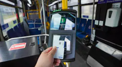 A rider taps a Visa card against a fare reader aboard a Milwaukee County Transit System bus. The reader's screen displays 'Open Payment – $2.75' with a green upward arrow, indicating a successful contactless transaction. The bus interior, with blue seating and yellow handrails, is visible in the background.