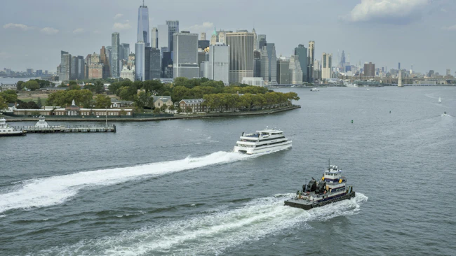 Two vessels cross New York Harbor with lower Manhattan's skyline and Governors Island in the background. A white passenger ferry cuts a wake in the middle distance while a smaller tugboat or work vessel passes in the foreground. One World Trade Center rises above the dense cluster of skyscrapers under a partly cloudy sky.