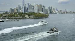 Two vessels cross New York Harbor with lower Manhattan's skyline and Governors Island in the background. A white passenger ferry cuts a wake in the middle distance while a smaller tugboat or work vessel passes in the foreground. One World Trade Center rises above the dense cluster of skyscrapers under a partly cloudy sky.