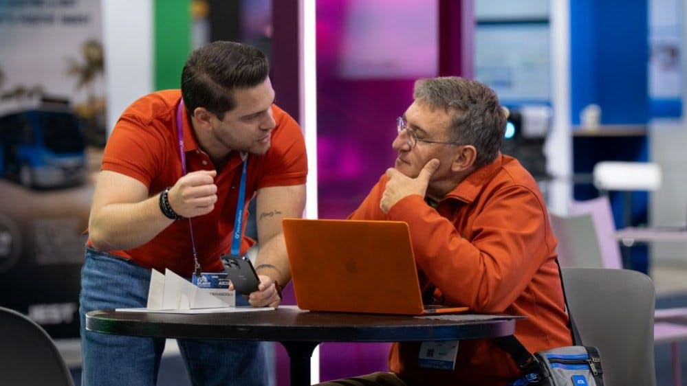 Two conference attendees confer at a small round table on a trade show floor. One person leans in while gesturing, the other listens thoughtfully with a hand to their chin, a laptop open between them. Colorful exhibit booths are visible in the background.