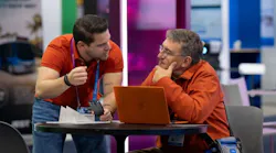 Two conference attendees confer at a small round table on a trade show floor. One person leans in while gesturing, the other listens thoughtfully with a hand to their chin, a laptop open between them. Colorful exhibit booths are visible in the background.