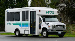 A white WTA (Whatcom Transportation Authority) paratransit/cutaway bus, fleet number 24718, built on a Ford E-Series chassis with an Endera body, photographed in a three-quarter view in a parking area surrounded by evergreen trees. The vehicle features the WTA logo and a teal, green, and blue horizontal stripe livery, a wheelchair accessibility symbol near the front passenger door, and a Washington state license plate. The door is open, revealing yellow interior handrails.
