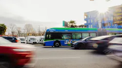 A TransLink RapidBus travels through a busy urban intersection in Metro Vancouver, surrounded by moving vehicles. The bus features the agency's signature green, blue and white livery. Construction of a new apartment building is visible in the background.