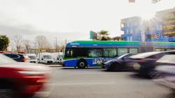 A TransLink RapidBus travels through a busy urban intersection in Metro Vancouver, surrounded by moving vehicles. The bus features the agency's signature green, blue and white livery. Construction of a new apartment building is visible in the background. A TransLink RapidBus travels through a busy urban intersection in Metro Vancouver, surrounded by moving vehicles. The bus features the agency's signature green, blue and white livery. Construction of a new apartment building is visible in the background.