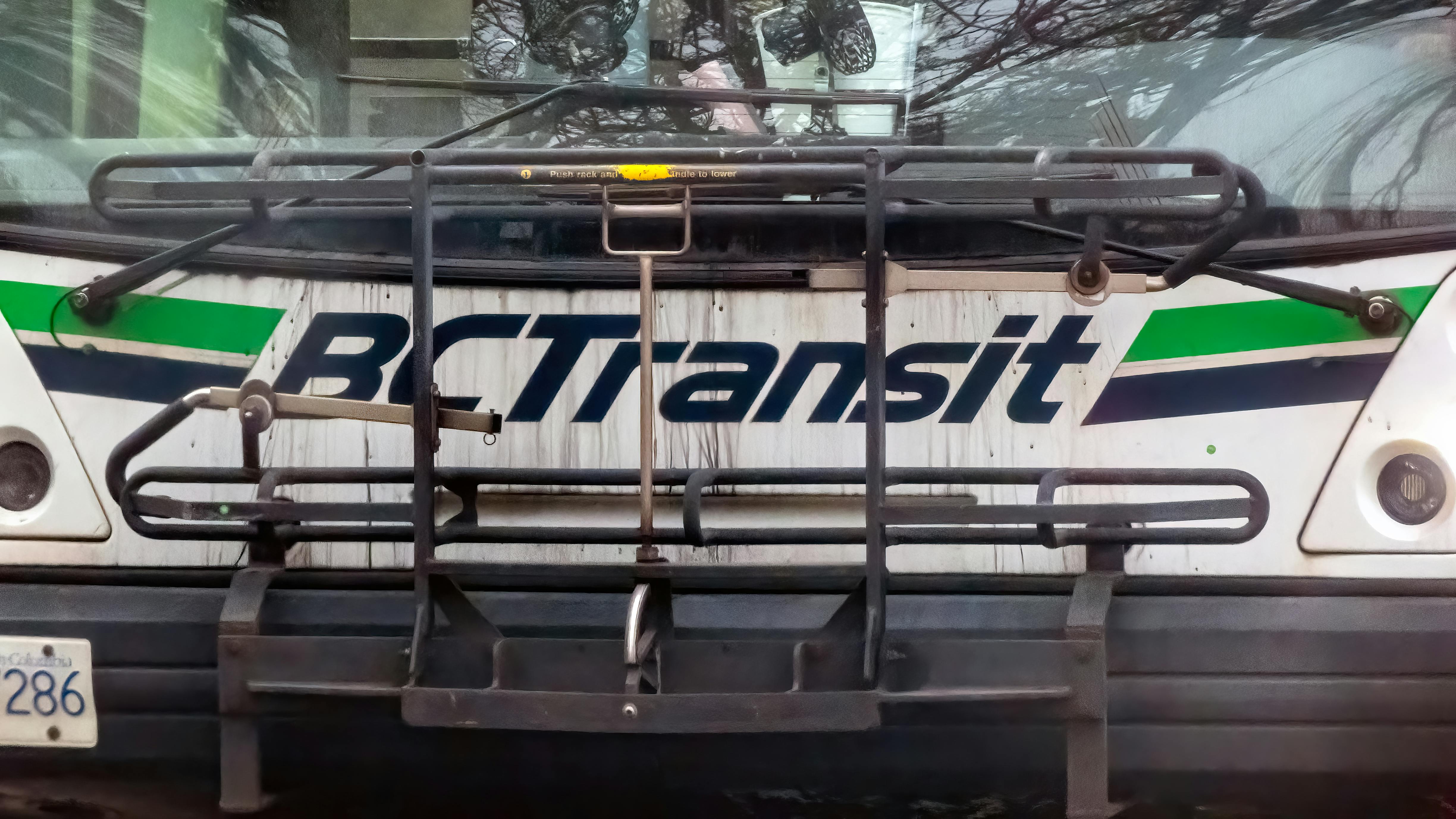 A close-up front view of a BC Transit bus, showing the agency's white livery with green and dark blue stripe. A bicycle rack is mounted on the front of the bus. Bare tree branches are reflected in the windshield.