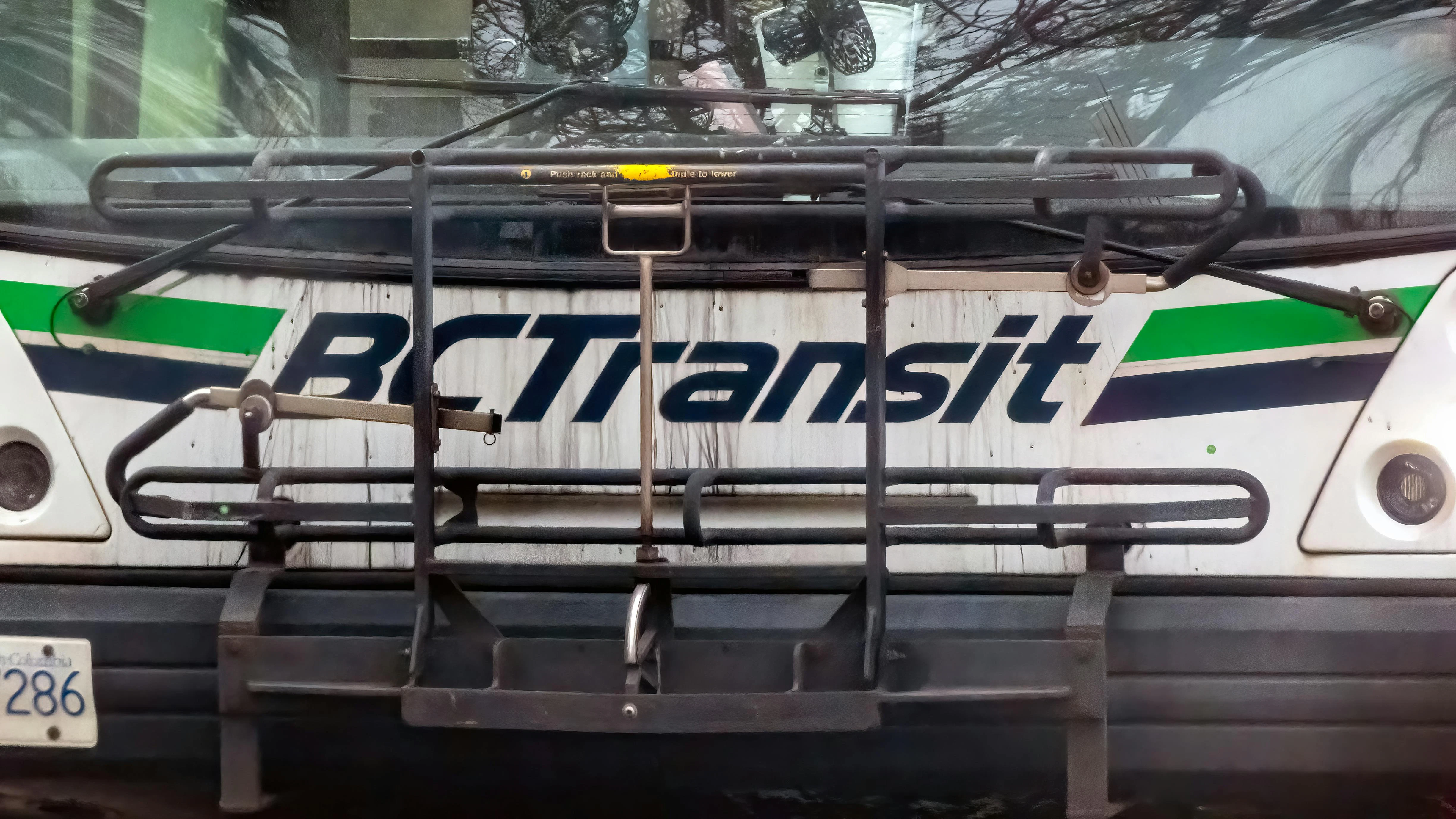 A close-up front view of a BC Transit bus, showing the agency's white livery with green and dark blue stripe. A bicycle rack is mounted on the front of the bus. Bare tree branches are reflected in the windshield.