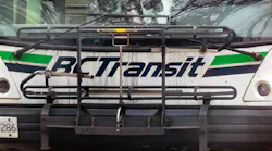 A close-up front view of a BC Transit bus, showing the agency's white livery with green and dark blue stripe. A bicycle rack is mounted on the front of the bus. Bare tree branches are reflected in the windshield.