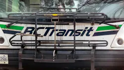 A close-up front view of a BC Transit bus, showing the agency's white livery with green and dark blue stripe. A bicycle rack is mounted on the front of the bus. Bare tree branches are reflected in the windshield. A close-up front view of a BC Transit bus, showing the agency's white livery with green and dark blue stripe. A bicycle rack is mounted on the front of the bus. Bare tree branches are reflected in the windshield.