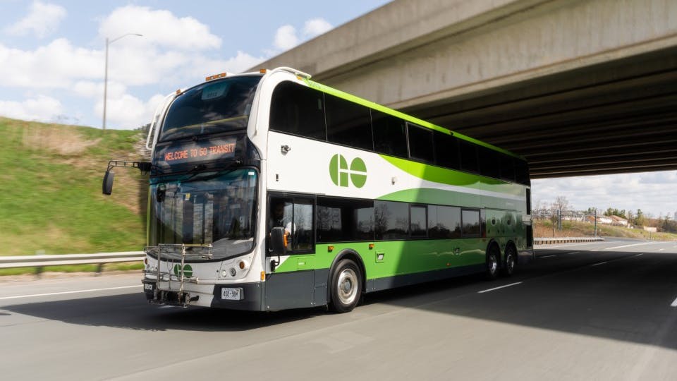 A GO Transit double-decker bus travels along a highway beneath an overpass. The destination sign reads 'Welcome to GO Transit.' The bus features the agency's white and green livery with the GO logo on its upper front panel.
