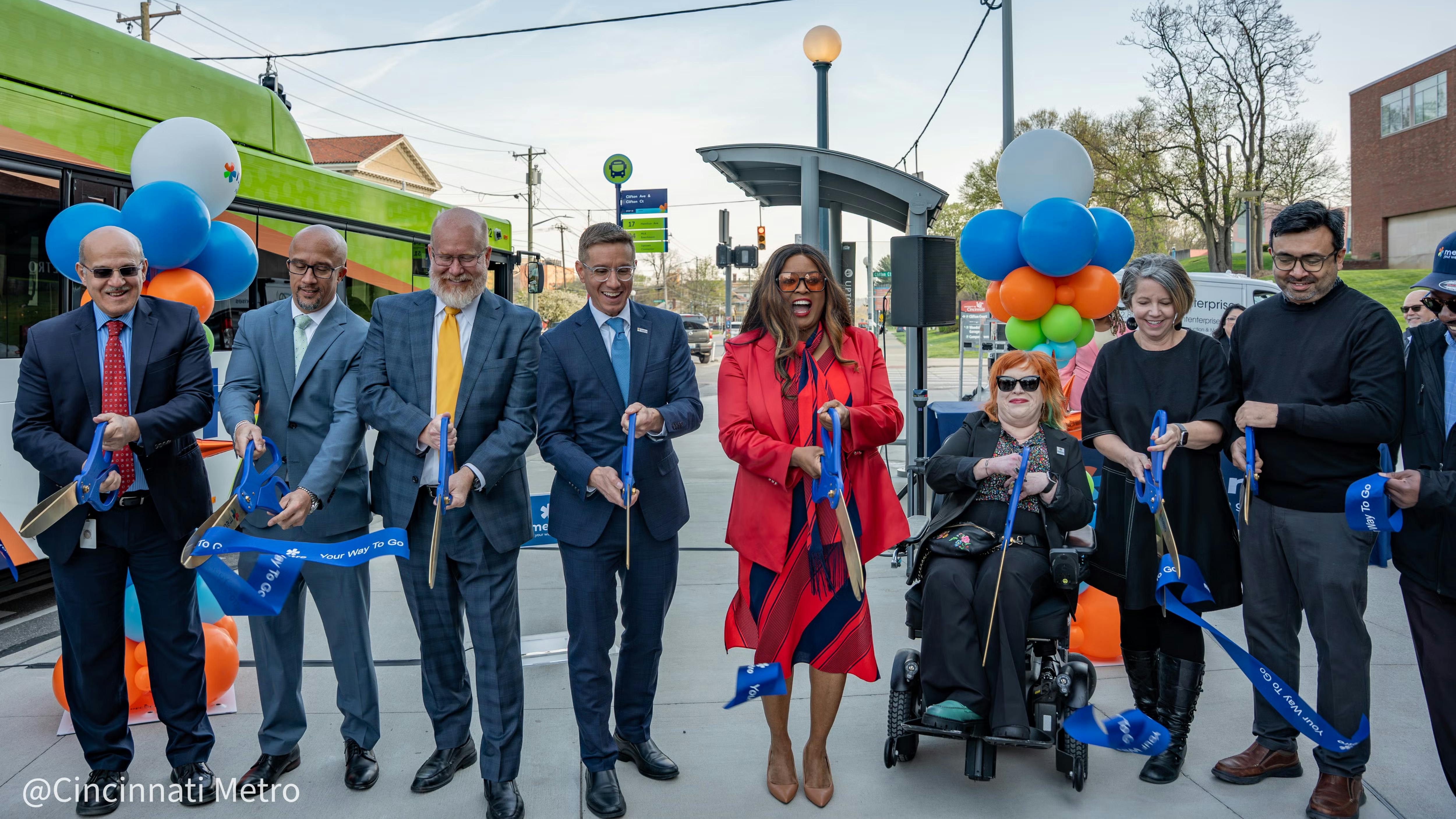 A group of approximately eight officials and community members participate in a ribbon-cutting ceremony at a Cincinnati Metro bus stop, holding blue ceremonial scissors and a blue ribbon reading 'Your Way To Go.' The group stands in front of the Clifton Avenue and Clifton Court stop sign and a green Cincinnati Metro bus. Blue, orange and green balloon clusters flank the group. One participant uses a power wheelchair. The photo is credited to Cincinnati Metro.