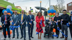 A group of approximately eight officials and community members participate in a ribbon-cutting ceremony at a Cincinnati Metro bus stop, holding blue ceremonial scissors and a blue ribbon reading 'Your Way To Go.' The group stands in front of the Clifton Avenue and Clifton Court stop sign and a green Cincinnati Metro bus. Blue, orange and green balloon clusters flank the group. One participant uses a power wheelchair. The photo is credited to Cincinnati Metro.