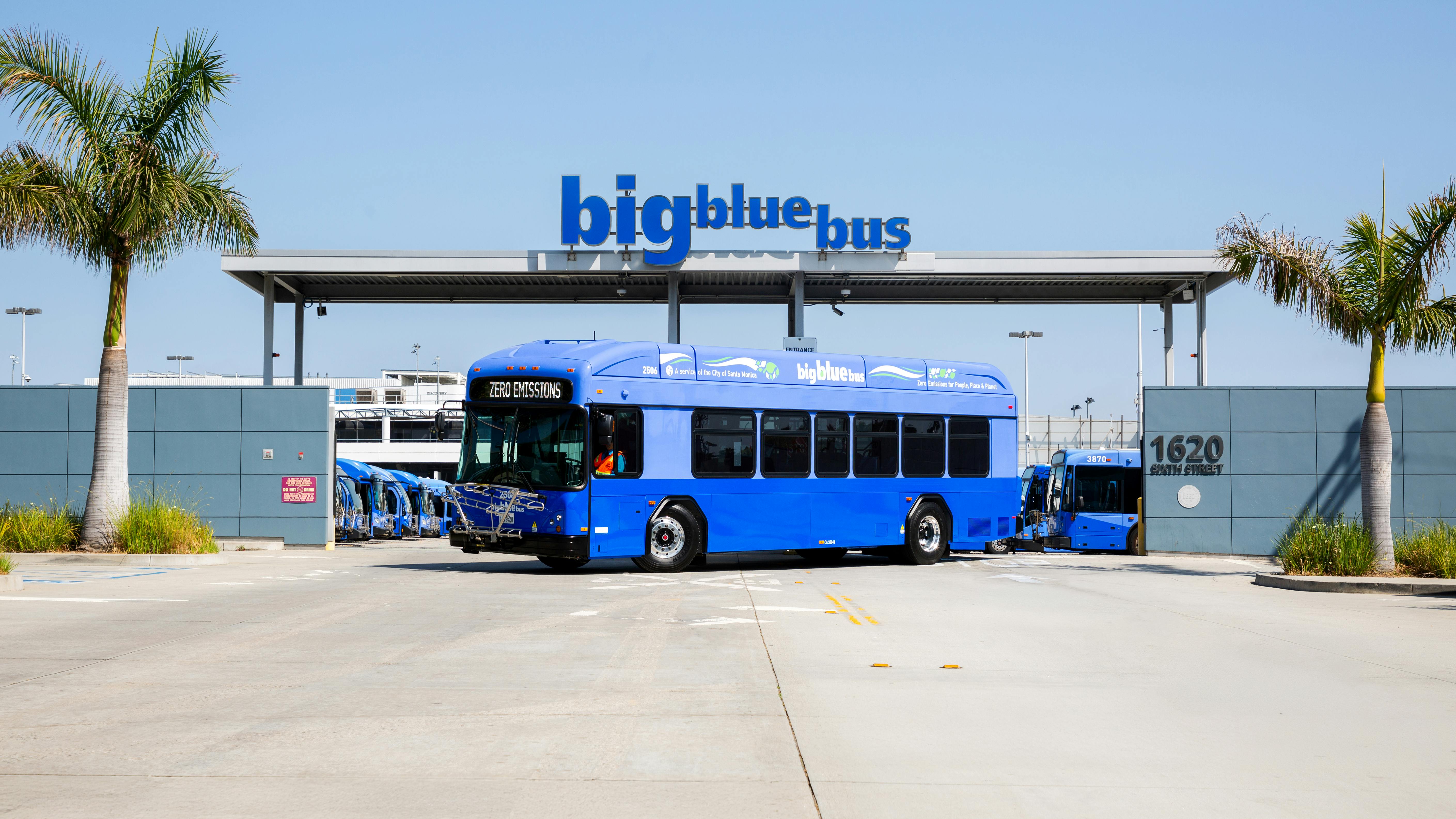 A Big Blue Bus battery-electric bus displaying 'Zero Emissions' on its destination sign sits parked in front of the agency's maintenance and operations facility at 1620 Sixth Street in Santa Monica, Calif., beneath a large 'big blue bus' sign mounted on a canopy at the facility entrance. Additional blue buses are visible parked inside the yard. Palm trees flank the entrance on both sides under a clear blue sky.