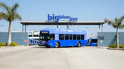 A Big Blue Bus battery-electric bus displaying 'Zero Emissions' on its destination sign sits parked in front of the agency's maintenance and operations facility at 1620 Sixth Street in Santa Monica, Calif., beneath a large 'big blue bus' sign mounted on a canopy at the facility entrance. Additional blue buses are visible parked inside the yard. Palm trees flank the entrance on both sides under a clear blue sky.
