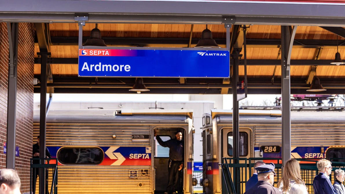 A blue station sign reading 'Ardmore' hangs from the canopy of the renovated Ardmore station on the Southeastern Pennsylvania Transportation Authority (SEPTA) regional rail network, with both the SEPTA and Amtrak logos displayed on the sign. Below, a SEPTA regional rail car is stopped at the platform with its doors open and a crew member visible in the doorway. A small group of passengers and officials gather on the platform beneath the canopy, which features warm wood paneling and pendant lights. A red metal roof is visible above.