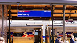 A blue station sign reading 'Ardmore' hangs from the canopy of the renovated Ardmore station on the Southeastern Pennsylvania Transportation Authority (SEPTA) regional rail network, with both the SEPTA and Amtrak logos displayed on the sign. Below, a SEPTA regional rail car is stopped at the platform with its doors open and a crew member visible in the doorway. A small group of passengers and officials gather on the platform beneath the canopy, which features warm wood paneling and pendant lights. A red metal roof is visible above.