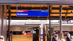 A blue station sign reading 'Ardmore' hangs from the canopy of the renovated Ardmore station on the Southeastern Pennsylvania Transportation Authority (SEPTA) regional rail network, with both the SEPTA and Amtrak logos displayed on the sign. Below, a SEPTA regional rail car is stopped at the platform with its doors open and a crew member visible in the doorway. A small group of passengers and officials gather on the platform beneath the canopy, which features warm wood paneling and pendant lights. A red metal roof is visible above. A blue station sign reading 'Ardmore' hangs from the canopy of the renovated Ardmore station on the Southeastern Pennsylvania Transportation Authority (SEPTA) regional rail network, with both the SEPTA and Amtrak logos displayed on the sign. Below, a SEPTA regional rail car is stopped at the platform with its doors open and a crew member visible in the doorway. A small group of passengers and officials gather on the platform beneath the canopy, which features warm wood paneling and pendant lights. A red metal roof is visible above.